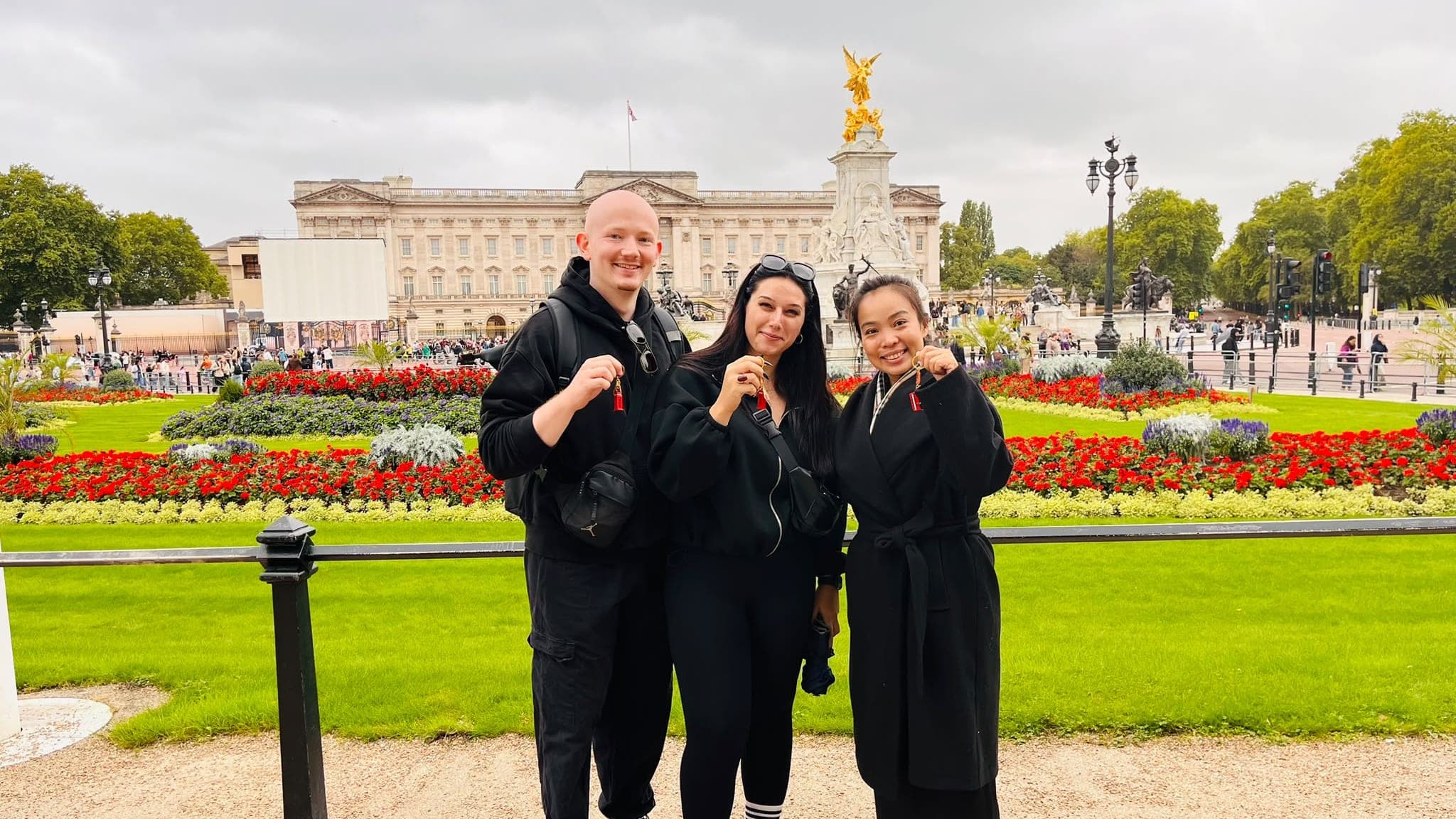 Three BeExplorers guests holding up their keyring souvenirs in front of Buckingham Palace